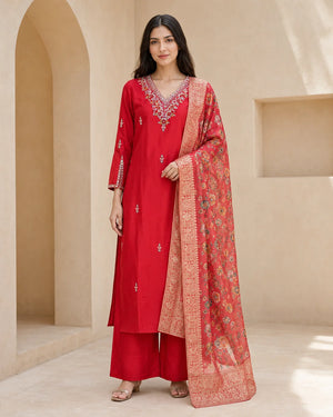 A woman models a traditional red Indian salwar kameez suit with a heavily embroidered red dupatta, set against a warm beige architectural background with arches.