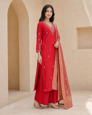 A woman models a traditional red Indian salwar kameez suit with silver embroidery and a peach dupatta, set against a beige architectural background with arches.