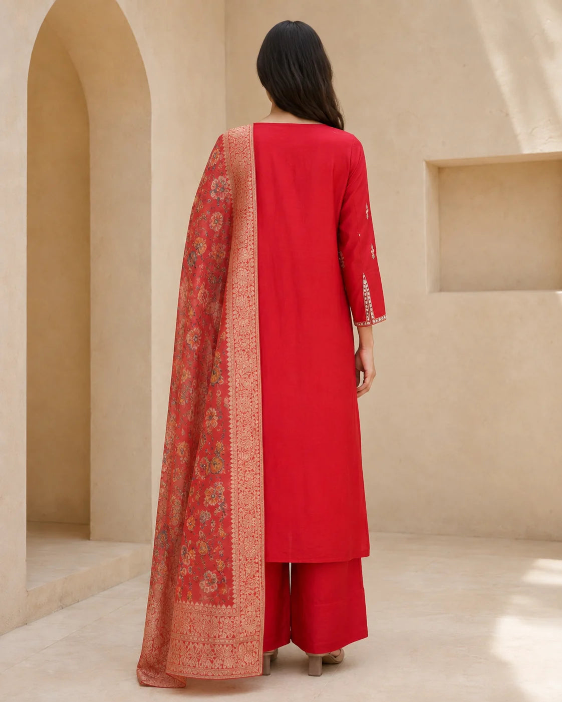 A woman viewed from behind wearing a bright red Indian ethnic outfit with matching trousers and a patterned dupatta draped over her left shoulder, set against a beige architectural background.