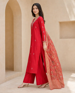 A woman modeling a red Indian ethnic suit with matching red pants and a peach-orange patterned dupatta, standing in a beige architectural setting.