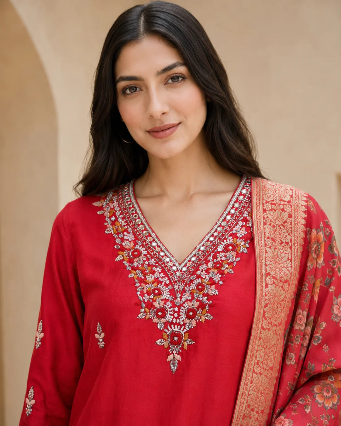 A woman wearing a red traditional embroidered kurta with a matching patterned dupatta, set against a warm beige architectural background.