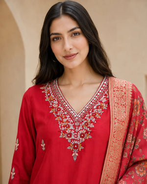 A woman wearing a red traditional embroidered kurta with a matching patterned dupatta, set against a warm beige architectural background.