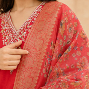 A close-up of a person wearing a red and coral floral embroidered traditional garment, showing the embroidered neckline and a patterned draped fabric with a hand pointing to the textile detail.