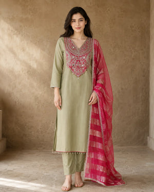 A woman models a traditional Indian salwar kameez suit set. The kurta is a light sage green with elaborate pink and silver embroidery around the neckline, paired with matching sage green trousers and a bright pink dupatta featuring a metallic gold border.
