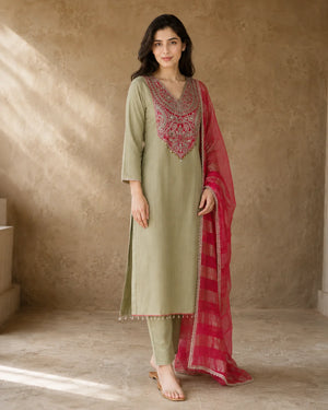 A woman models a traditional Indian salwar kameez suit set in a warm, textured studio setting. The set includes an olive green kurta with a heavily embroidered pink and gold neckline, matching olive green trousers, and a bright pink dupatta with gold stripes.