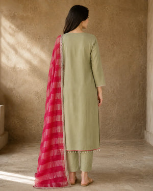 A woman stands with her back to the camera wearing a light sage green three-piece salwar kameez suit with a bright pink and gold striped dupatta, set against a textured beige wall.