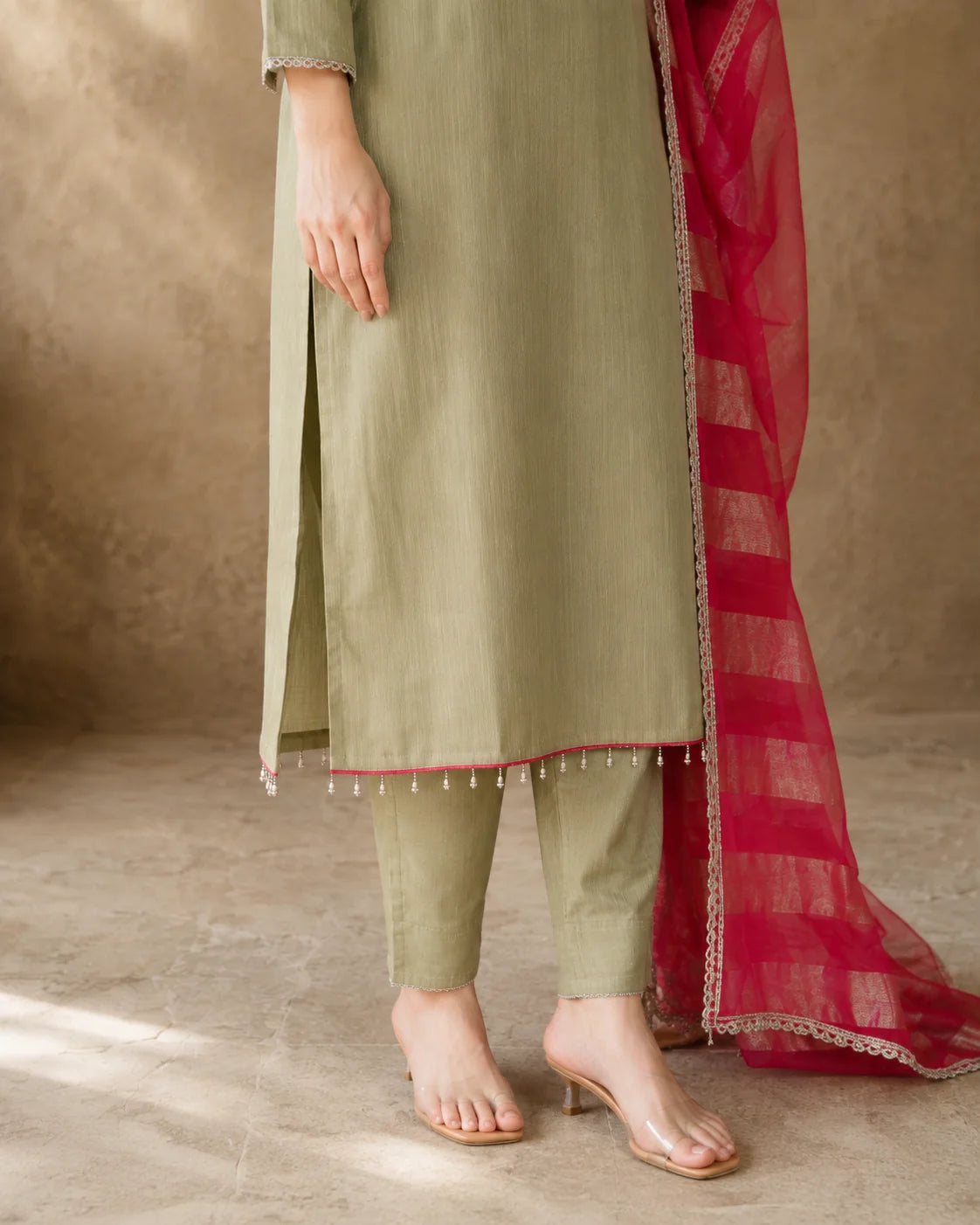 A person wearing an olive green traditional outfit with matching pants and a bright red sheer dupatta, standing against a textured beige wall and floor.