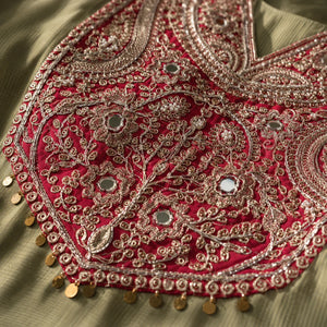 Close-up of an ornate red embroidered neckline with metallic gold threadwork, floral patterns, and small mirrored embellishments on a light olive fabric.
