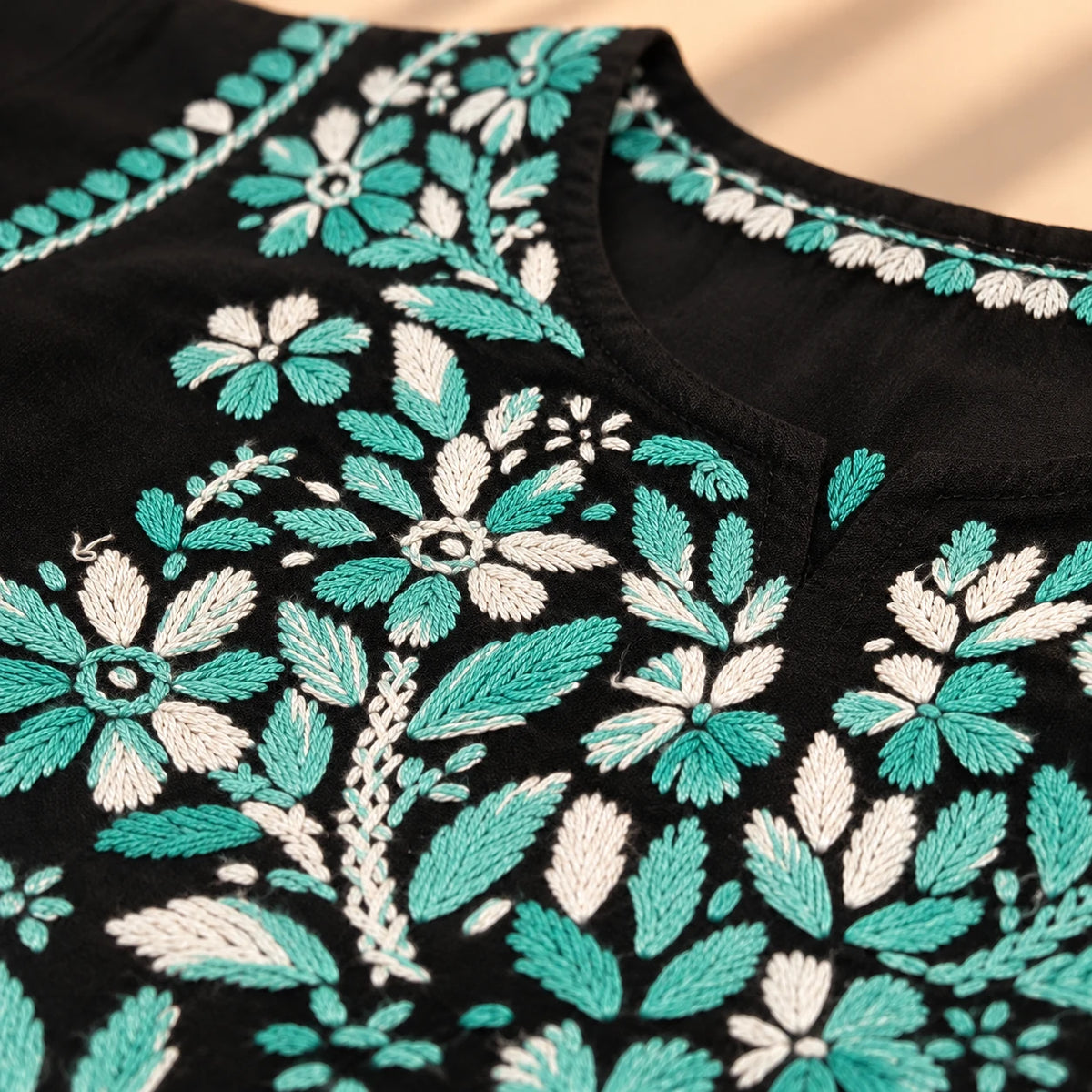 Close-up of a black embroidered garment featuring intricate floral and leaf patterns in teal and off-white thread.