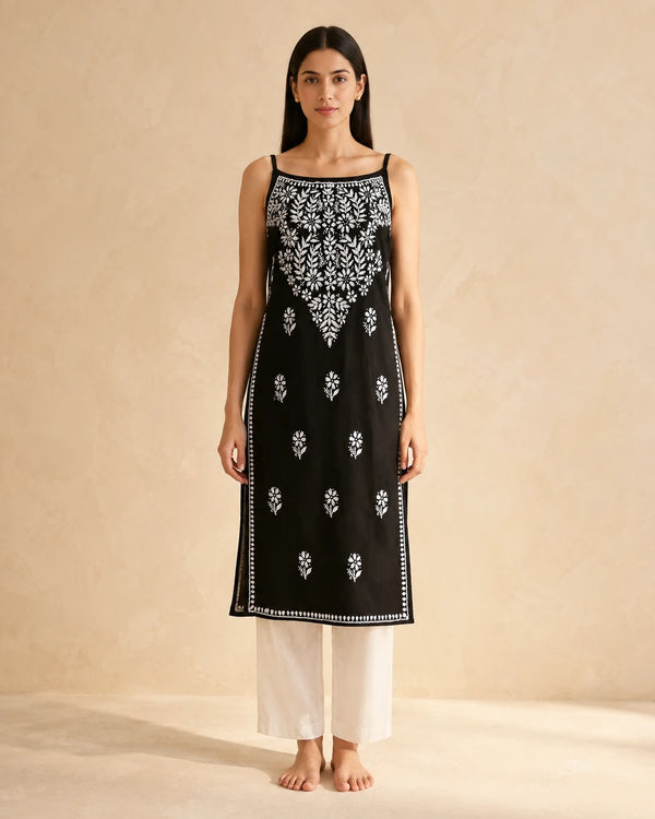 A woman stands barefoot against a warm beige textured wall, wearing a sleeveless black kurta with white floral embroidery and white pants.