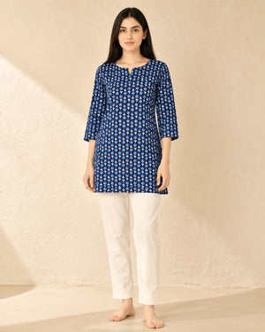 A woman stands barefoot against a textured beige wall wearing a blue and white patterned tunic top with three-quarter sleeves and white pants.