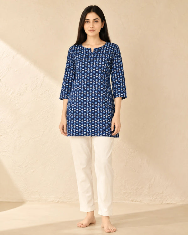 A woman stands barefoot against a textured beige wall wearing a blue and white patterned tunic top with three-quarter sleeves and white pants.