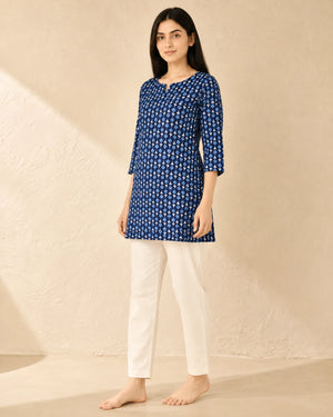 A woman stands barefoot against a textured beige wall wearing a blue printed kurta with white three-quarter sleeves and white pants.