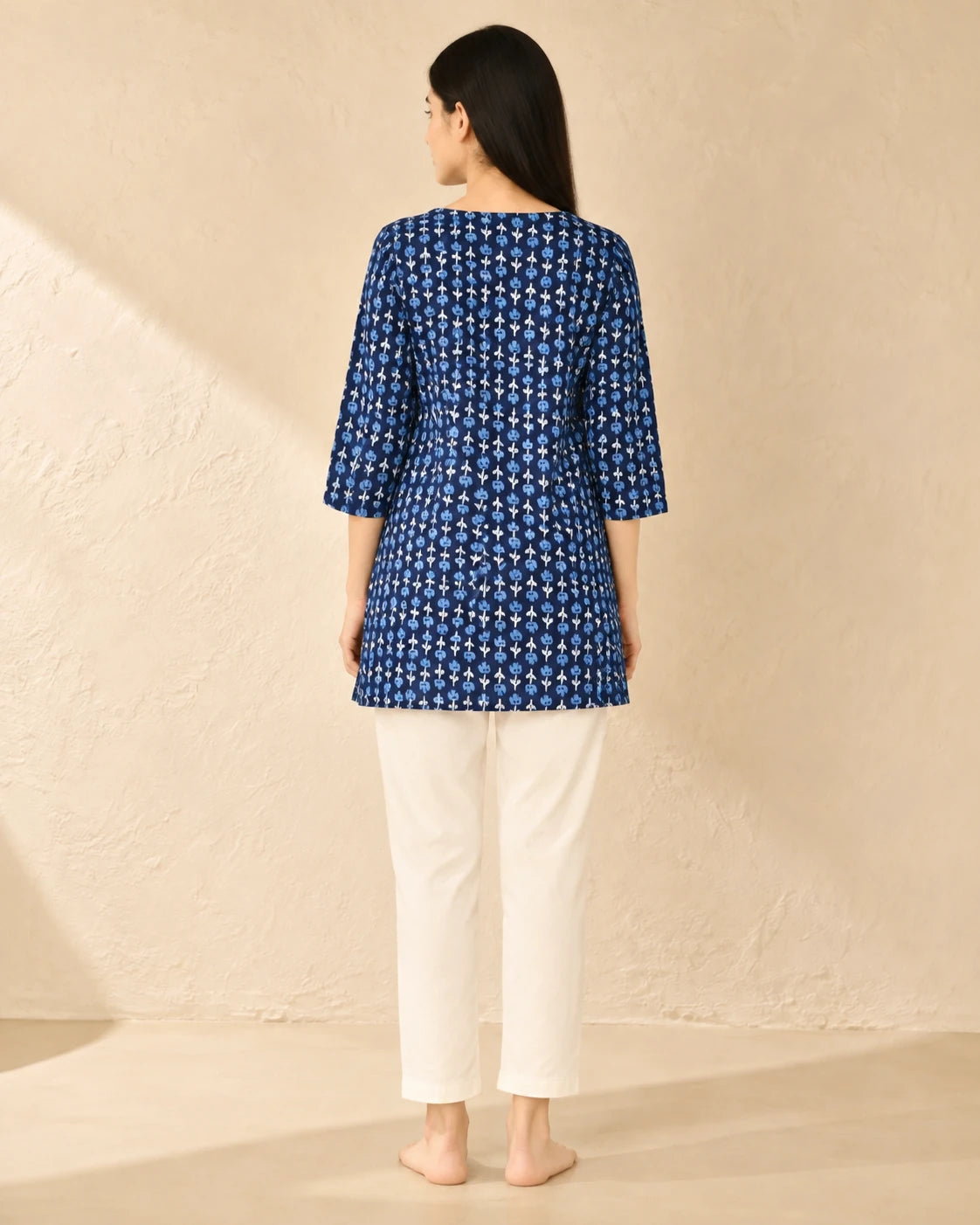 A woman viewed from the back wearing a blue three-quarter sleeve kurta with a white floral pattern, paired with white cropped pants, standing against a textured beige wall.