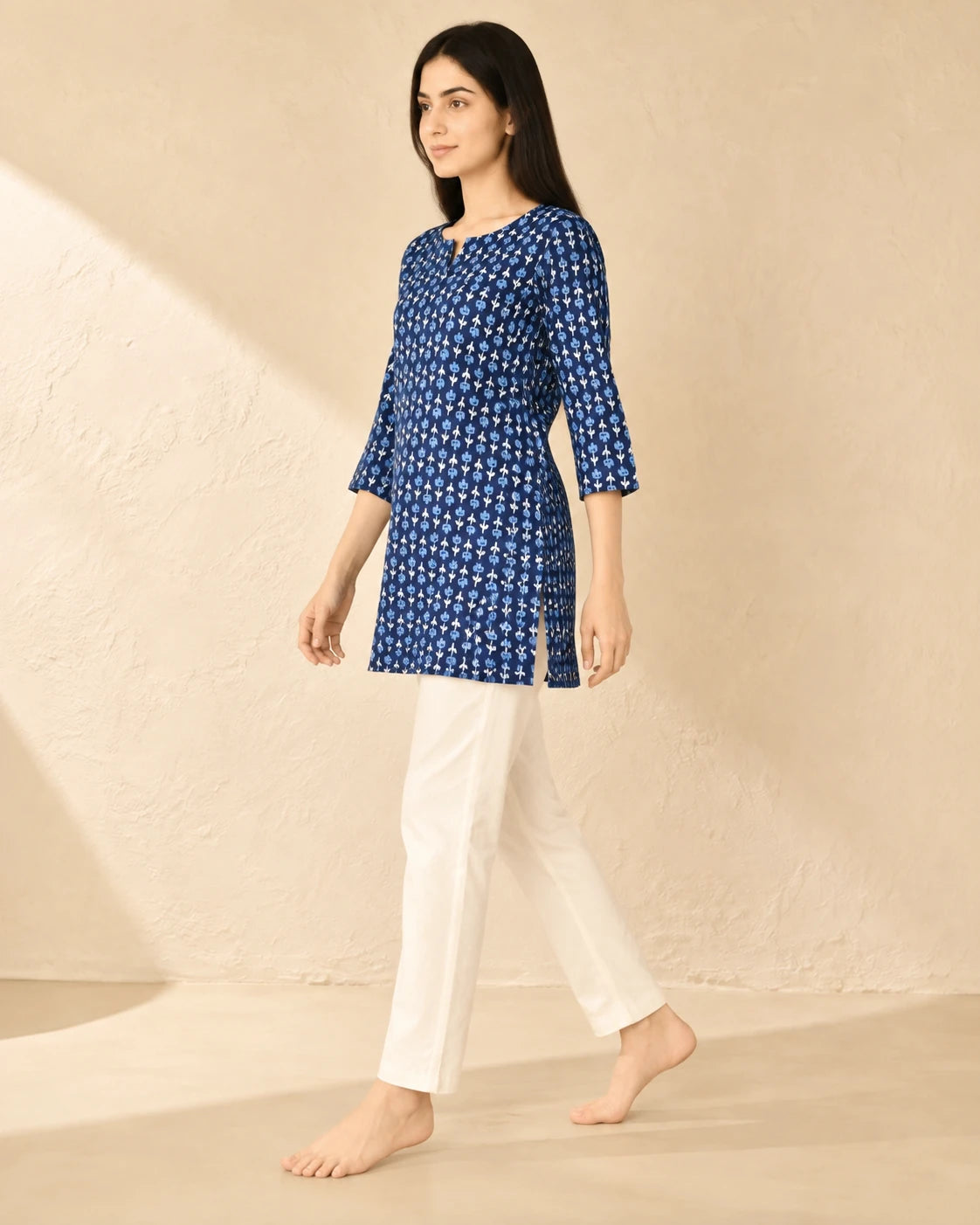 A woman modeling a blue and white patterned kurta with three-quarter sleeves, paired with white trousers, standing against a textured beige wall.
