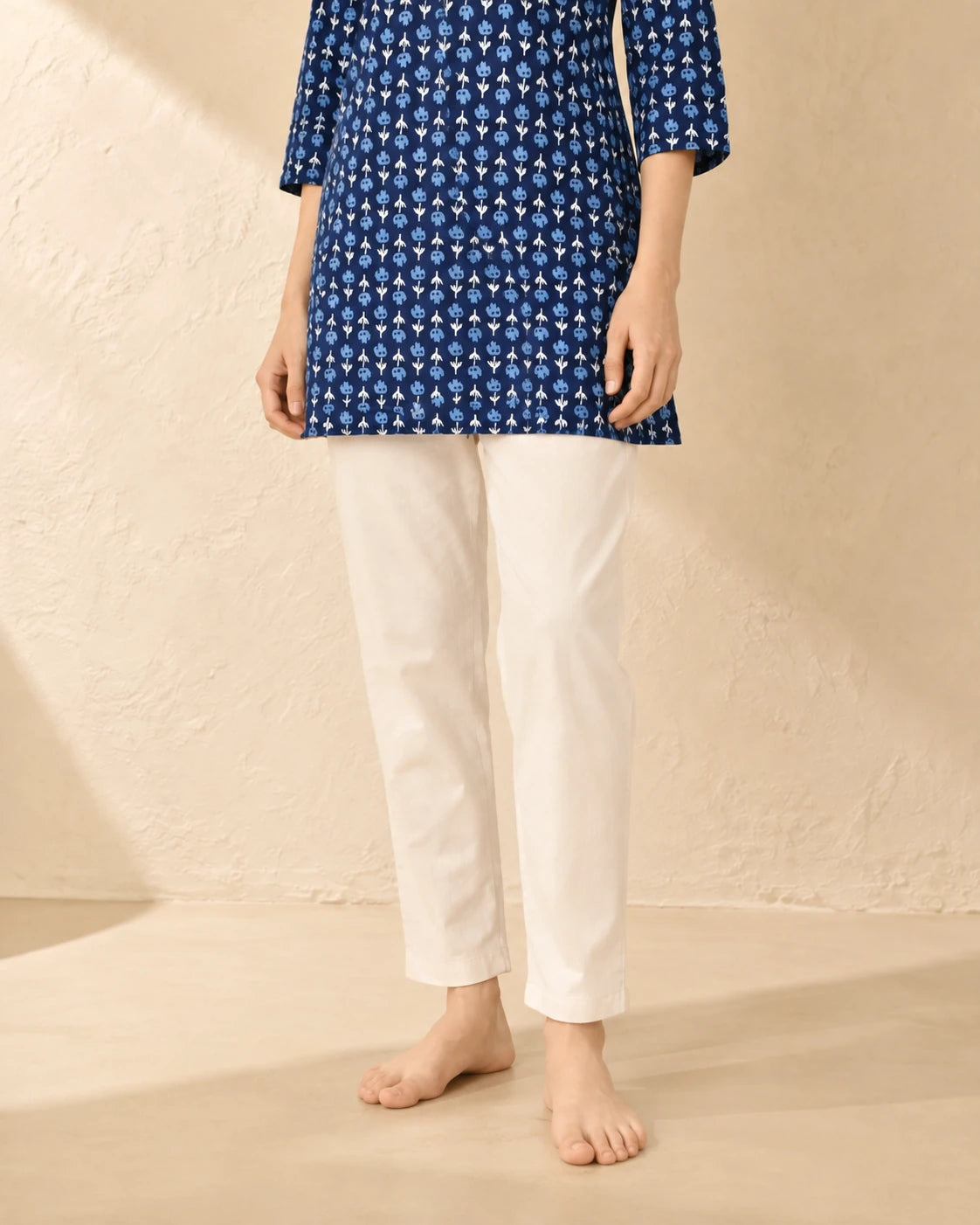 A model wearing a navy blue printed tunic top with white three-quarter sleeves and white cropped trousers, standing barefoot against a textured beige wall.