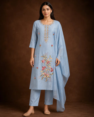 A woman modeling a light blue traditional Indian salwar kameez suit with floral embroidery on the front, matching trousers, and a sheer draped scarf, set against a dark brown studio background.