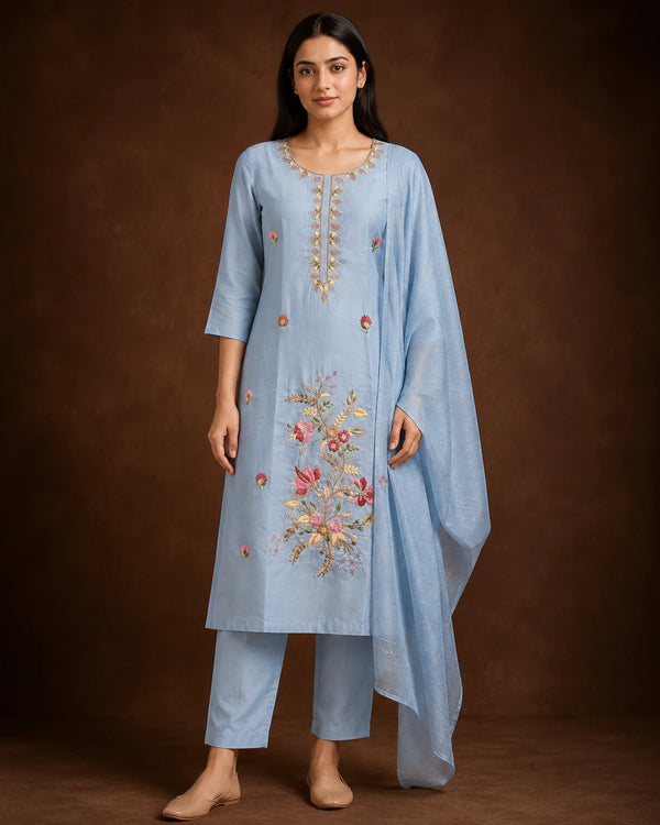 A woman modeling a light blue traditional Indian salwar kameez suit with floral embroidery on the front, matching trousers, and a sheer draped scarf, set against a dark brown studio background.