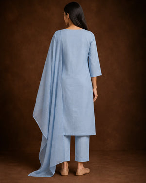 A woman shown from the back wearing a light blue three-piece traditional outfit with a long draped top, matching cropped pants, and a long matching scarf against a brown studio backdrop.