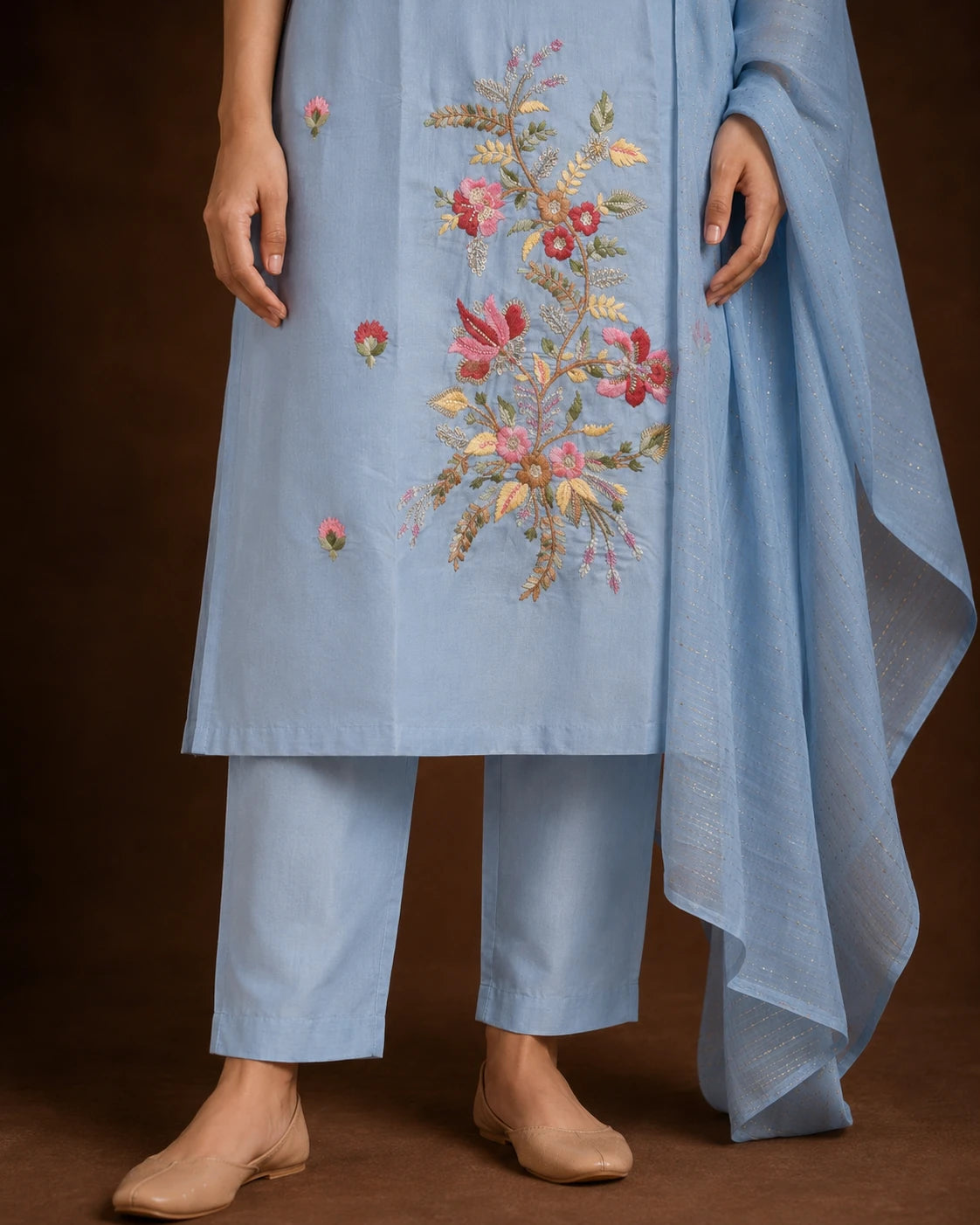 A model wearing a light blue traditional outfit with floral embroidery on the front, matching pants, and a draped dupatta, photographed against a dark brown studio background.