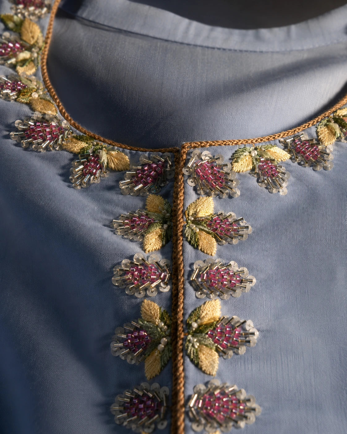Close-up of a blue-gray embroidered garment neckline with metallic gold thread, pink beaded floral motifs, and green leaf details.
