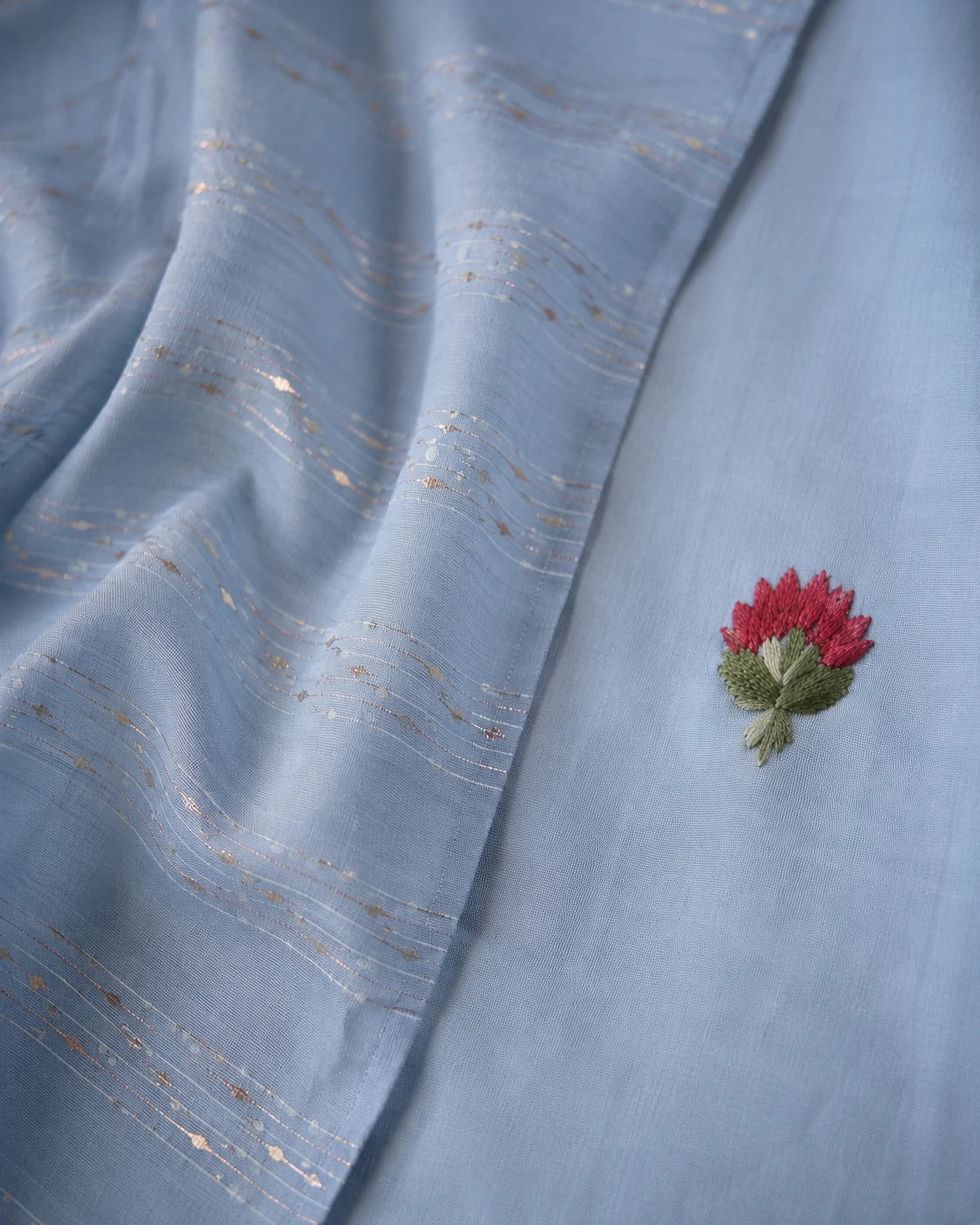 A close-up of a light blue fabric, likely a dupatta or saree pallu, featuring delicate gold zari embroidered wavy lines and a small red and green floral applique.