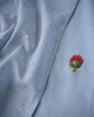 A close-up of a light blue fabric, likely a dupatta or saree pallu, featuring delicate gold zari embroidered wavy lines and a small red and green floral applique.
