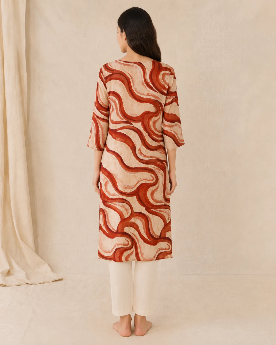 A woman stands with her back to the camera wearing a beige tunic with a rust-red wavy abstract pattern and white pants, set against a neutral cream backdrop.