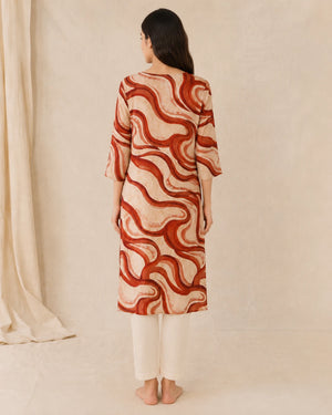 A woman stands with her back to the camera wearing a beige tunic with a rust-red wavy abstract pattern and white pants, set against a neutral cream backdrop.