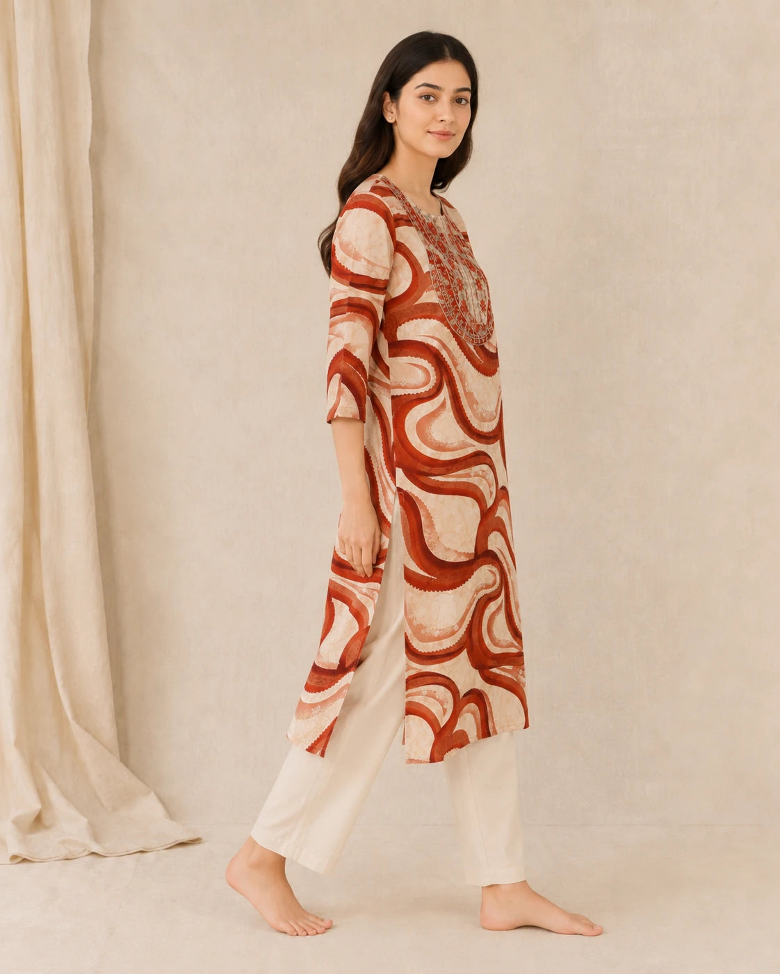 A woman modeling a knee-length kurta with three-quarter sleeves, featuring an abstract wavy pattern in rust red and cream, paired with cream-colored pants, set against a neutral beige backdrop.