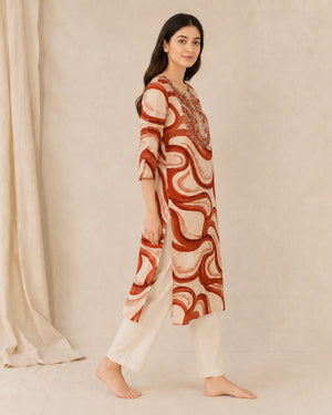 A woman modeling a knee-length kurta with three-quarter sleeves, featuring an abstract wavy pattern in rust red and cream, paired with cream-colored pants, set against a neutral beige backdrop.