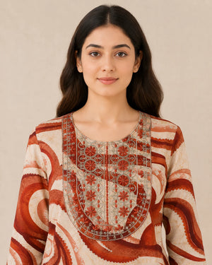A woman wearing a cream-colored tunic top with rust-red and brown abstract swirl patterns and a detailed embroidered yoke, set against a plain beige background.