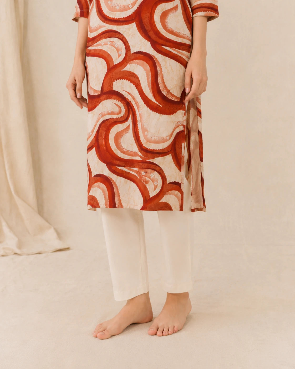 A person wearing a cream-colored kurta with a bold red and orange wavy abstract pattern, paired with white pants, standing barefoot against a warm beige background.