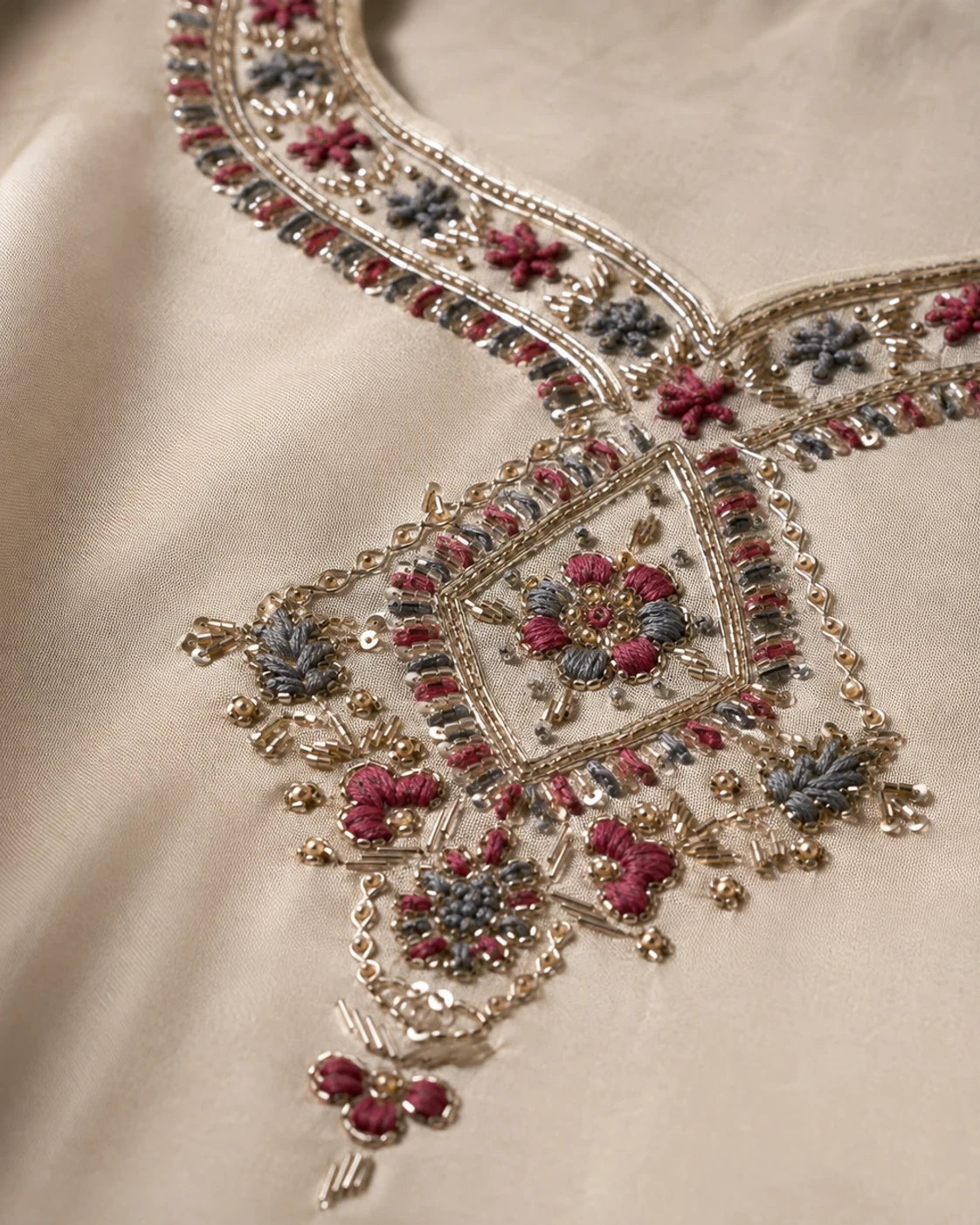 Close-up of an ornate beige fabric garment featuring intricate gold embroidery with pink, blue, and silver floral motifs.