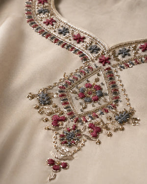 Close-up of an ornate beige fabric garment featuring intricate gold embroidery with pink, blue, and silver floral motifs.