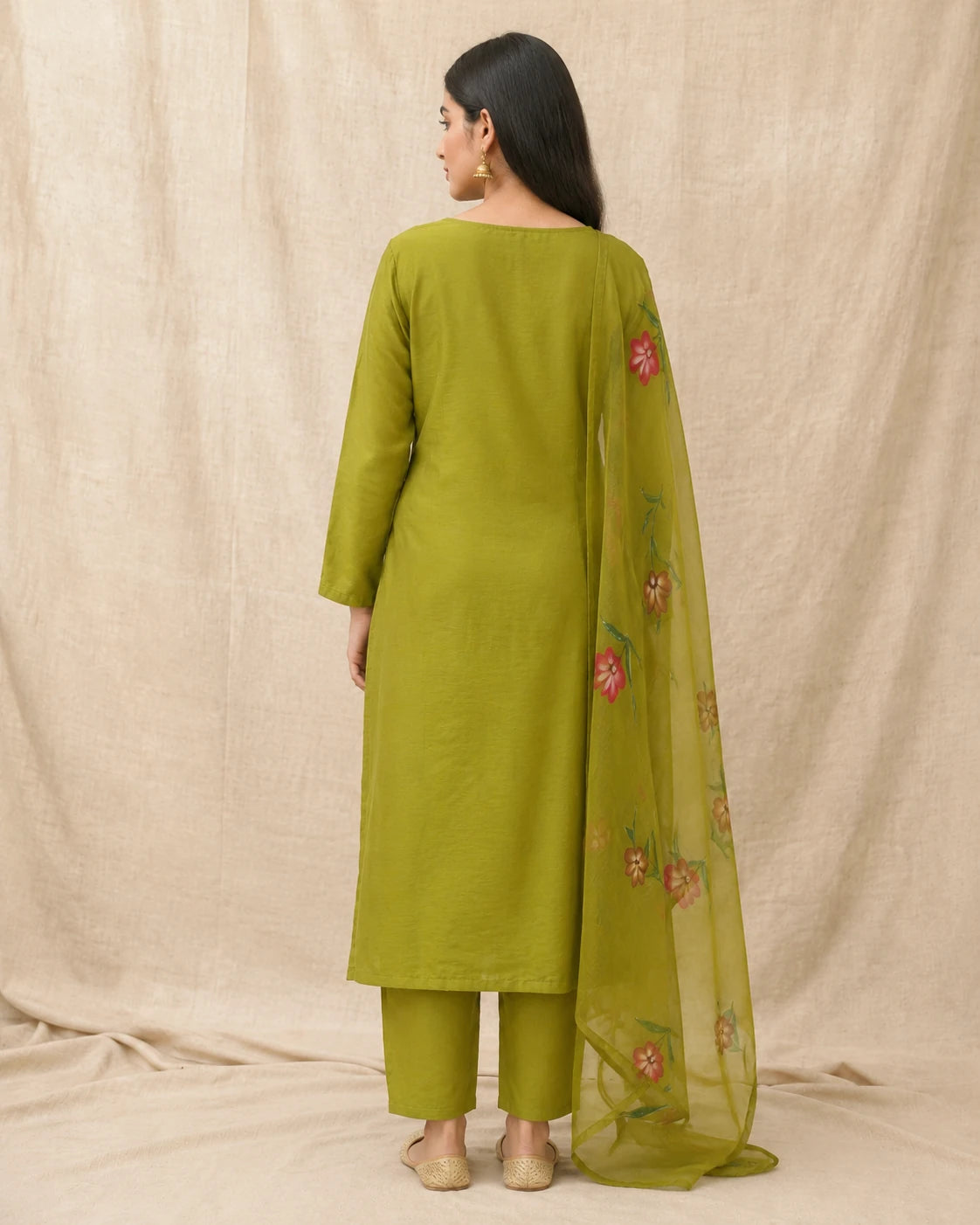 A woman shown from the back wearing a sage green kurta and matching pants with a sheer green dupatta featuring pink floral embroidery, set against a neutral beige fabric backdrop.