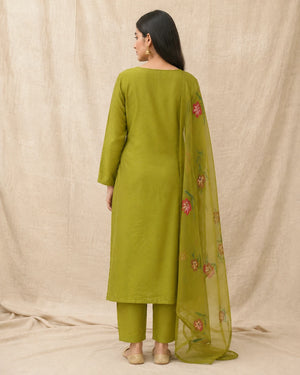 A woman shown from the back wearing a sage green kurta and matching pants with a sheer green dupatta featuring pink floral embroidery, set against a neutral beige fabric backdrop.