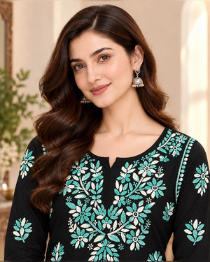 A woman wearing a black long-sleeve kurta with intricate teal and white floral embroidery, styled with silver jhumka earrings in a warm indoor setting.
