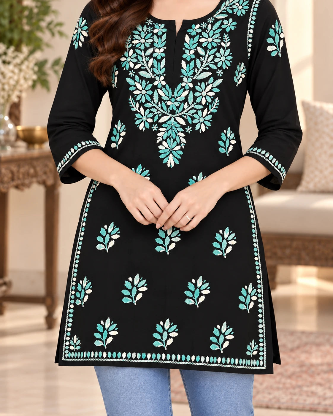 A woman wearing a black kurta with turquoise and white floral embroidery, paired with light blue jeans, in a softly lit indoor setting.