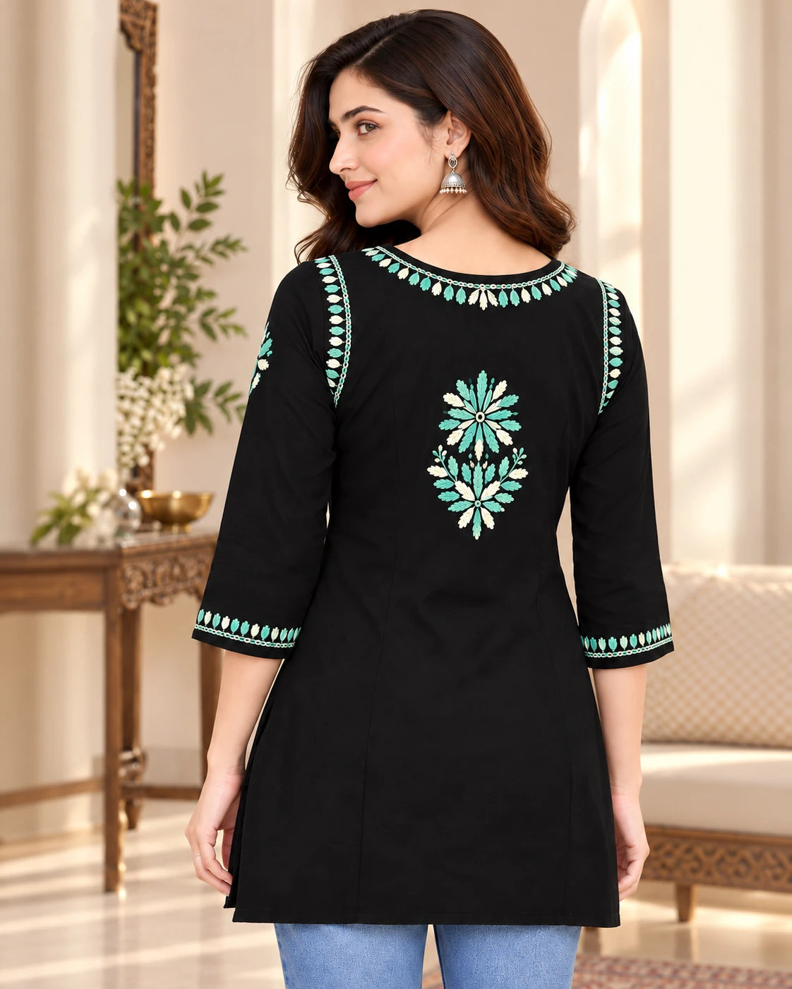 A woman modeling a black three-quarter sleeve kurta with turquoise and white floral embroidery on the back and sleeve cuffs, set against an elegant indoor background.