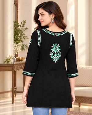A woman modeling a black three-quarter sleeve kurta with turquoise and white floral embroidery on the back and sleeve cuffs, set against an elegant indoor background.