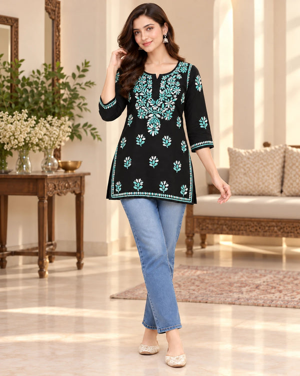 A woman models a black three-quarter sleeve kurta with intricate teal and white floral embroidery, paired with light blue jeans and beige flat shoes in a beautifully decorated indoor living room setting.