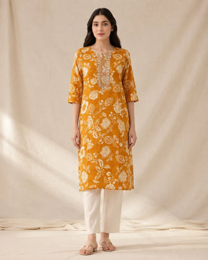A woman stands against a draped cream fabric backdrop wearing a mustard yellow kurta with a white floral print and white pants.