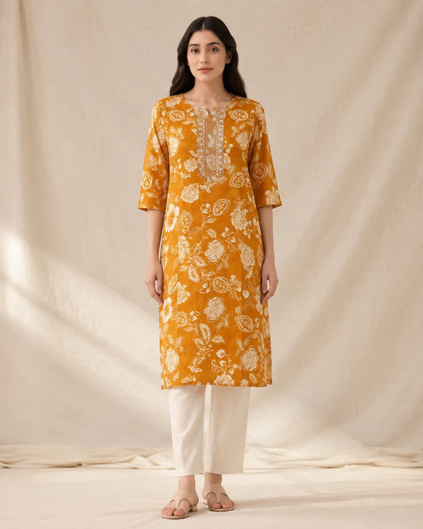 A woman stands against a draped cream fabric backdrop wearing a mustard yellow kurta with a white floral print and white pants.