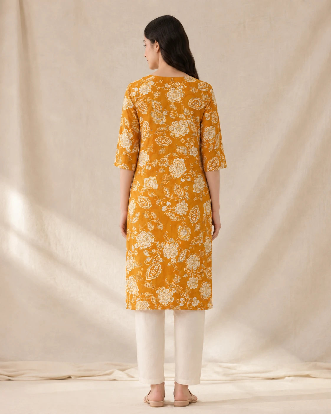 A woman viewed from behind wearing a mustard yellow kurta with a white floral block print, paired with off-white trousers, standing against a draped beige fabric backdrop.