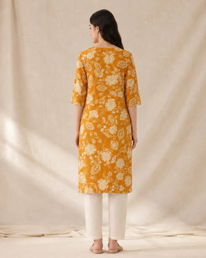 A woman viewed from behind wearing a mustard yellow kurta with a white floral block print, paired with off-white trousers, standing against a draped beige fabric backdrop.