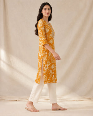 A woman modeling a mustard yellow kurta with a white floral print, paired with off-white pants and beige sandals, standing against a draped beige fabric backdrop.