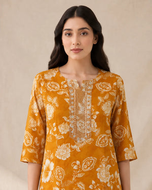 A woman modeling a mustard yellow kurta with a cream floral block print and a silver embroidered neckline, set against a neutral beige background.