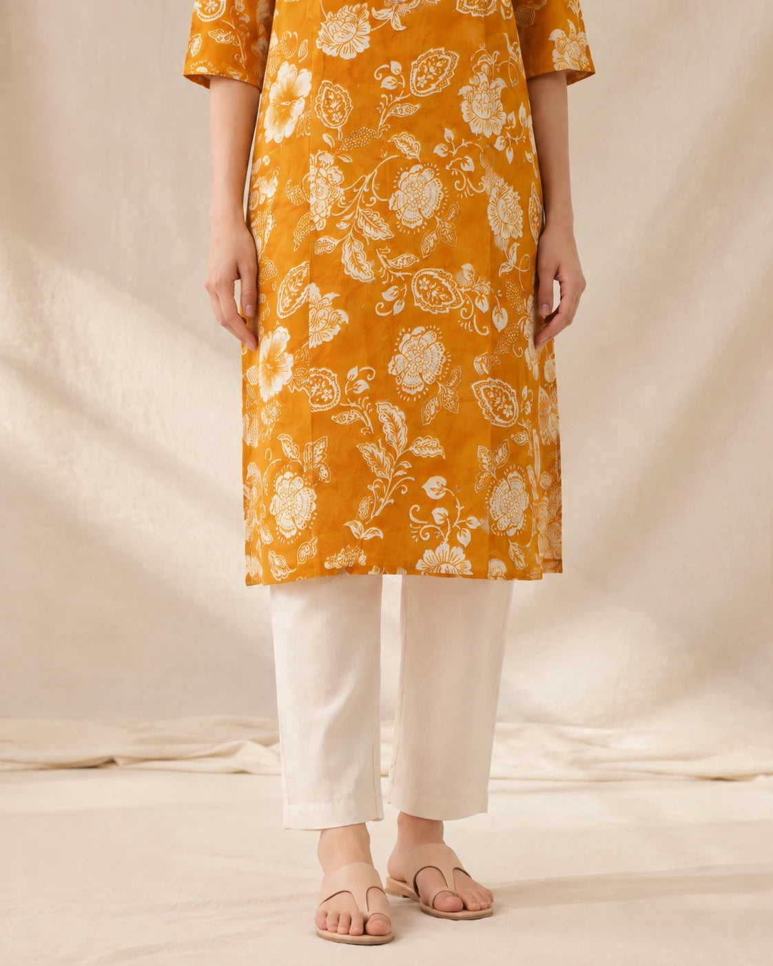 A person wearing a mustard yellow kurta with a white floral print, paired with off-white cropped pants and flat sandals, standing against a draped cream fabric backdrop.