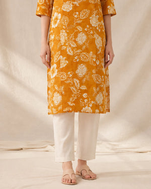 A person wearing a mustard yellow kurta with a white floral print, paired with off-white cropped pants and flat sandals, standing against a draped cream fabric backdrop.